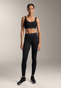 Czarny sportowy crop top i legginsy o wysokim stanie, obydwa wykonane z gładkiego materiału. Modelka nosi czarne trampki, neutralne tło.