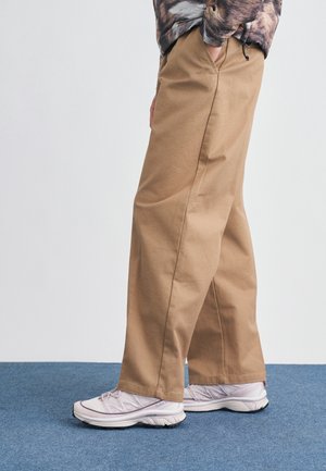 Person iført beige vidde bukser og hvide sneakers står på en blå måtte med hænderne i lommerne.