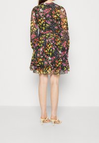 Robe florale noire avec de longues manches transparentes, présentant des motifs de fleurs colorés. Ourlet volanté et ceinture à nouer soulignant la taille. Portée avec des talons jaunes.