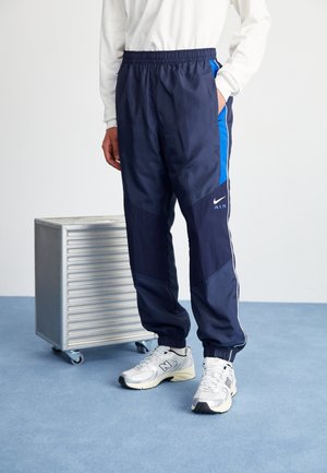Personne portant un pantalon de survêtement Nike Air bleu marine avec des panneaux latéraux bleus et des baskets blanches New Balance, debout à côté d'une boîte métallique argentée sur roulettes.
