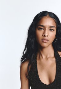 Czarna, bezrękawnikowa sukienka z głębokim dekoltem, o połyskującej teksturze; noszona przez modelkę o długich, falowanych włosach na jasnym tle.