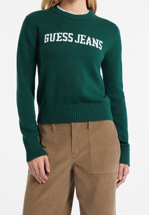 Kvinde iført en mørkegrøn sweater med teksten "GUESS JEANS" henover brystet, kombineret med lysebrune manchesterbukser.