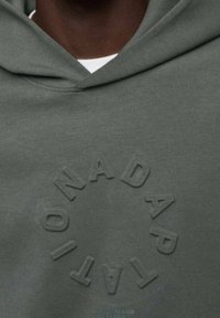 Gros plan d'un sweat à capuche vert foncé avec un texte circulaire en relief « ADAPTATION » autour de la poitrine.
