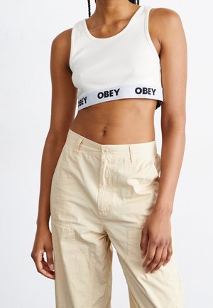 Witte geribbelde cropped top met een zwarte "OBEY" band, gecombineerd met lichtbeige hoge taillebroeken met zijzakken en een knoopsluiting.