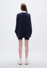 Μπλε σκούρο φαρδύ φούτερ με λευκό γιακά, μακριά μανίκια και άνετη εφαρμογή, συνδυασμένο με μαύρο σορτς και ουδέτερα αθλητικά παπούτσια.