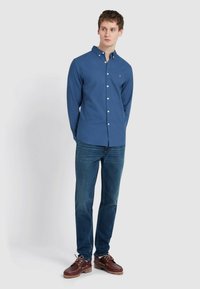 Blauwe langslijf hemdbloes met kraag, gecombineerd met donkerblauwe jeans en bruine veterschoenen. De stof lijkt glad met een minimale textuur.