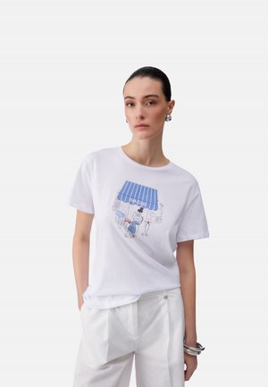 Femme aux cheveux plaqués en arrière portant des boucles d'oreilles argentées en forme de créoles, un T-shirt blanc avec une illustration de café bleue, un pantalon blanc et des bracelets argentés.