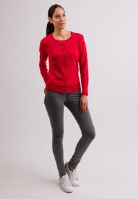 Czerwona kardigan z zapinaniem na guziki, okrągłym dekoltem, długimi rękawami i prążkowanym wykończeniem. Połączony z szarymi obcisłymi dżinsami i białymi trampkami.