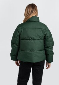 Giacca a vento verde con collo alto, texture trapuntata e vestibilità ampia, abbinata a pantaloni neri, vista da dietro.