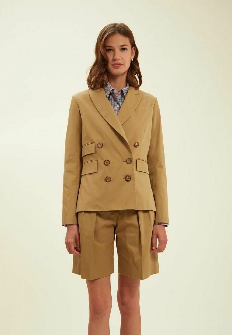 Blazer beige à double boutonnage avec des boutons ronds, deux poches avant et un short beige plissé. En dessous, une chemise à carreaux bleus est visible.