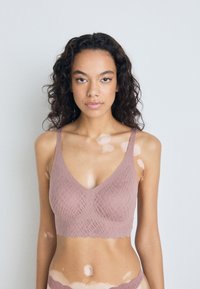 Mauve bešavna braletica s V-izrezom, teksturiranim dijamantnim uzorkom i valovitim rubom. Ima široke naramenice za potporu.