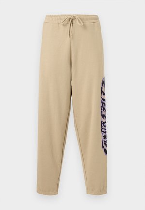 Béžové tepláky s elastickým pásom a sťahovacou šnúrkou, zúženými nohavicami a fialovým štýlizovaným grafickým textom na pravej strane.