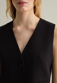 Gilet nero senza maniche con scollatura a V profonda, caratterizzato da due bottoni scuri e spalle leggermente imbottite; tessuto liscio e taglio aderente.