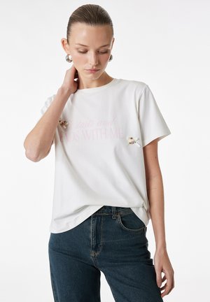 Vrouw met naar achteren gestyled haar draagt een wit T-shirt met kleine roze bloemenspeldjes en blauwe spijkerbroek, kijkt omlaag met hand op haar nek.