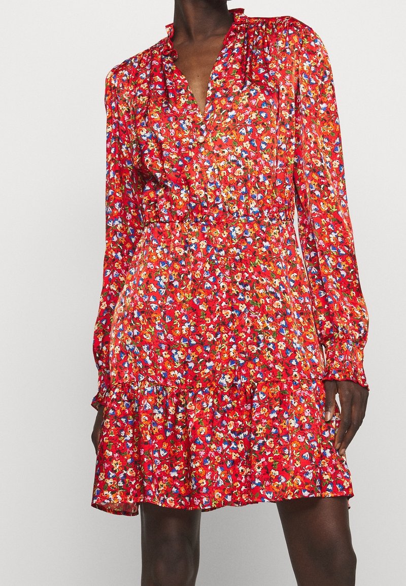 Robe courte rouge à manches longues avec imprimé floral multicolore, taille froncée, col en V boutonné et ourlet à volants sur une personne debout.