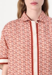 Korte mouwen shirt van perzik stof met een geometrisch patroon in rood en wit, met een contrasterende knoopsluiting en gestreepte mouwen.