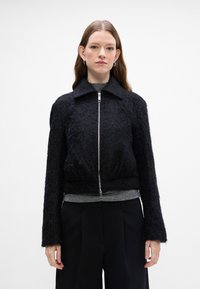 Čierna skrátená bunda vyrobená z textúrovaného materiálu; má predný zips a golier. Nosená cez šedý top a čierne široké nohavice.