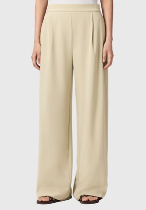 Beige broek met wijde pijpen en plooien aan de voorkant, gedragen door een persoon die staat, gecombineerd met donkere sandalen met open tenen.