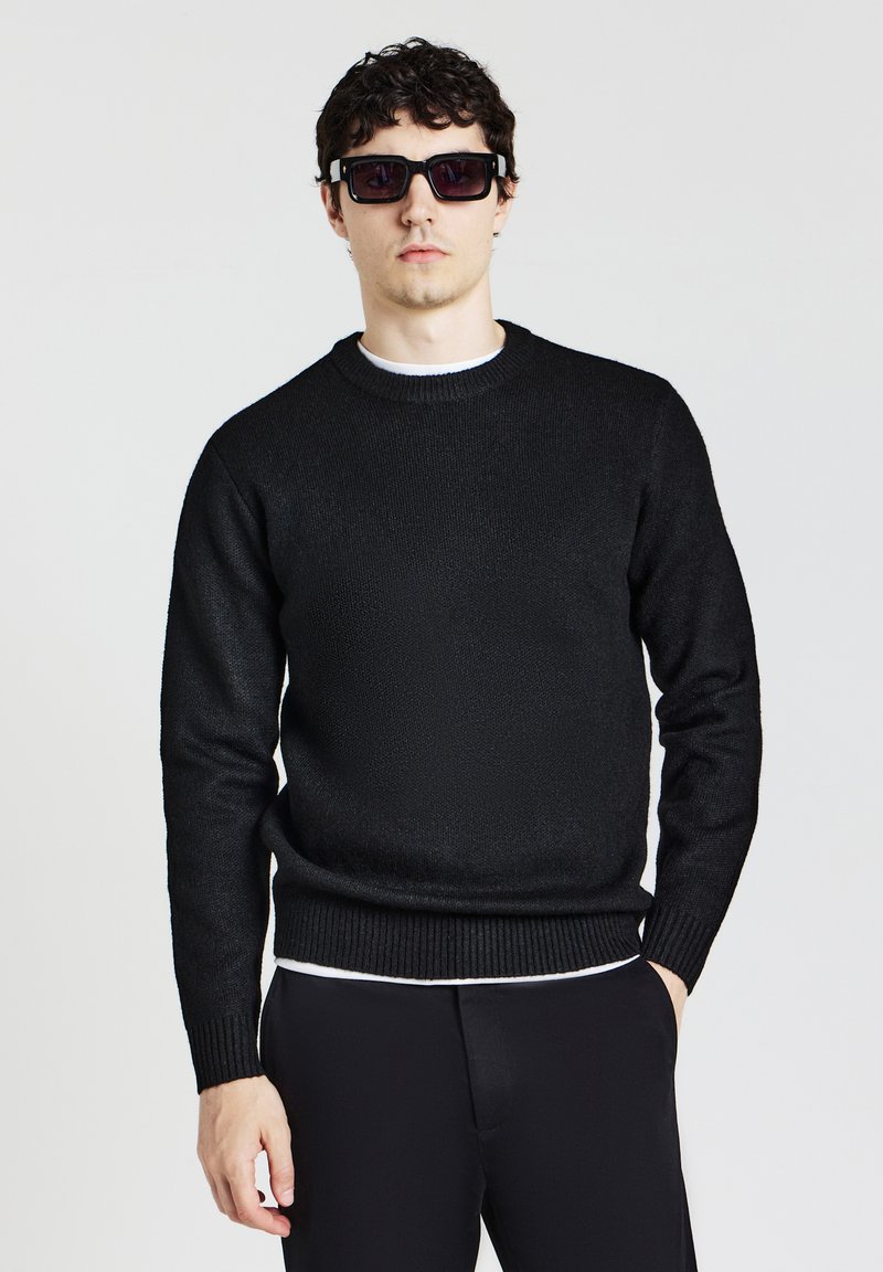 Schwarzer gestrickter Pullover mit rundem Ausschnitt, gerippten Bündchen und Saum. Modell trägt Sonnenbrillen, kombiniert mit einem weißen T-Shirt darunter.