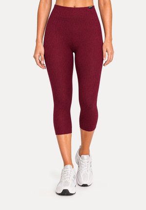 Femme portant des leggings bordeaux courts et des chaussures de sport blanches, debout devant un fond clair uni.