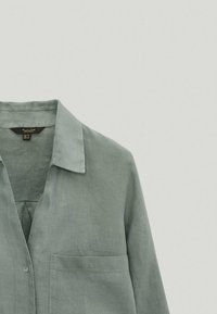 Chemise en lin vert clair avec col, poche poitrine et détails à boutons sur le devant. Texture lisse avec des accents de couture minimalistes.