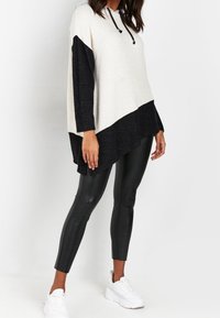 Kvinna klädd i en lös tvåfärgad svart och beige hoodie med svarta leggings i läderlook och vita sneakers, stående mot en vit bakgrund.