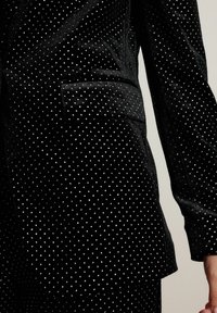 Sort fløjlsblazer med sølvfarvet pailletmønster, der har en enkelt brystlomme og detaljer i sidesømmen, struktureret snit med lange ærmer.