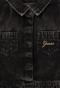 Melnas denima krekls ar izteiktu apkakli, divām krūšu kabatām un 'Guess' logo zelta izšuvumā, ar teksturētu auduma apdari.