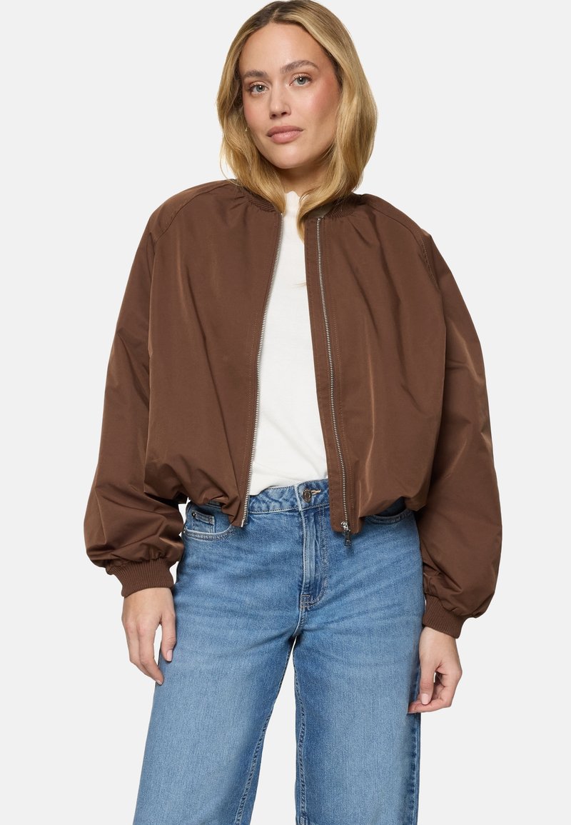 Femme aux cheveux blonds portant une veste bomber marron sur un haut blanc et un jean taille haute bleu, debout avec une posture détendue.