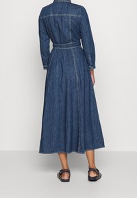 Woman wearing a long blue denim dress with belt detail at waist, buttoned cuffs, and black flat sandals, standing against a plain light background.