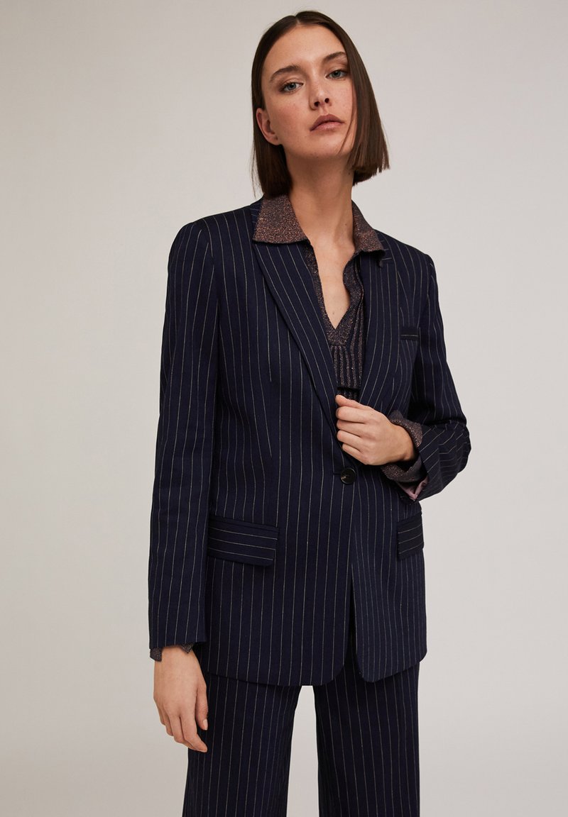 Blazer navy a righe sottili con revers a punta, chiusura a un bottone e tasche laterali. Include una camicia marrone texturizzata sotto.