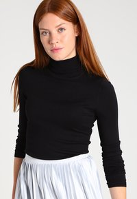 Schwarzer Rollkragenpullover aus dehnbarem Stoff, mit langen Ärmeln, figurbetonendem Design, kombiniert mit einem gefältelten, hellgrauen Rock.