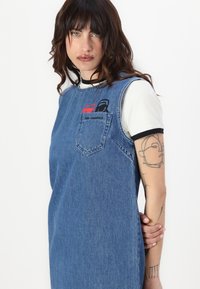 Vestido de ganga com design sem mangas, cor azul, bolso no peito e detalhes bordados em vermelho e preto. Camiseta simples branca com bordas pretas.