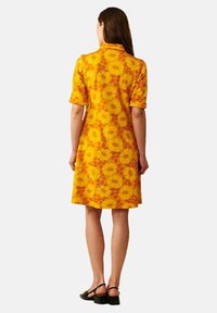 Keltainen lyhythihainen mekko, jossa on kukkakuosi, polvenmittainen, tasapintainen. Käytetty mustien sandaalien kanssa, nähty takaa.