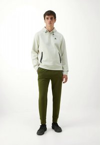 Sweat à capuche gris clair avec des accents noirs et des cordons de serrage, associé à un pantalon de jogging vert olive. Des chaussures de sport noires complètent la tenue.