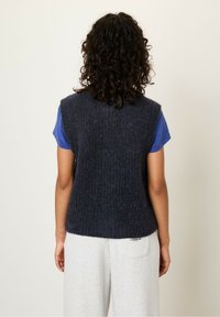 Persoon met krullend donker haar die een mouwloze donkerblauwe gebreide vest draagt over een blauwe t-shirt en lichtgrijze broek, met hun rug naar de kijker gekeerd.