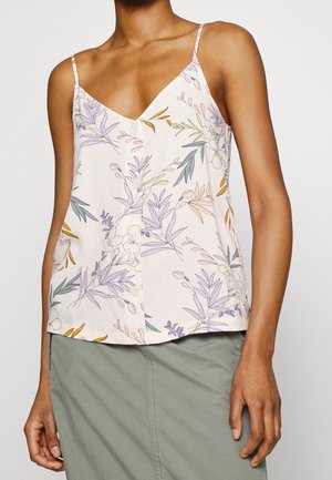 Femme portant un caraco blanc sans manches à imprimé floral violet et orange et une jupe gris clair sur un fond uni.