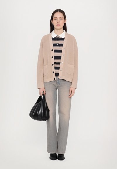 Μπεζ πλεκτό καρντάν με στρογγυλή λαιμόκοψη, κουμπάκια και τσέπες, φορεμένο πάνω από μια μαύρη και μπεζ ριγέ πόλο, συνδυασμένο με ανοιχτό τζιν και μια μαύρη τσάντα.