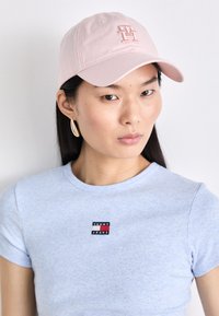 Cappellino da baseball rosa chiaro con logo ricamato, abbinato a una maglietta a coste blu chiaro con un piccolo patch di marca. Design semplice e moderno.