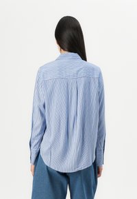 Camisa listrada azul e branca com um ajuste descontraído, frente com botões, mangas longas e bainha arredondada. O tecido parece leve e apresenta uma textura suave.