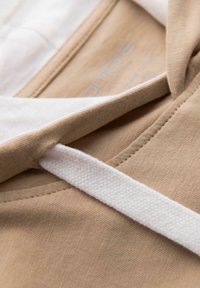 Detailní záběr na béžovou mikinu s bílou šňůrkou pro stažení provlečenou prošitým otvorem poblíž výstřihu.