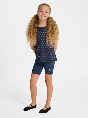 Smilende ung pige med langt krøllet hår i navy mønstret ærmeløs top, navy biker shorts og sorte balletflade stående med hænderne bag ryggen.