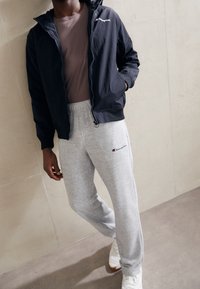 Námořnická lehká bunda s kapucí, světle hnědá přiléhavá košile, šedé tepláky a bílé tenisky s oranžovými akcenty. Viditelné logo Champion.