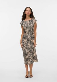 Femme portant une robe midi beige avec un motif de points noirs irréguliers, à manches courtes, et des sandales ouvertes noires, se tenant devant un fond blanc uni.