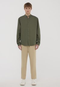 Camicia verde oliva con colletto alto e maniche lunghe, indossata con pantaloni beige e sneakers bianche, su uno sfondo neutro.