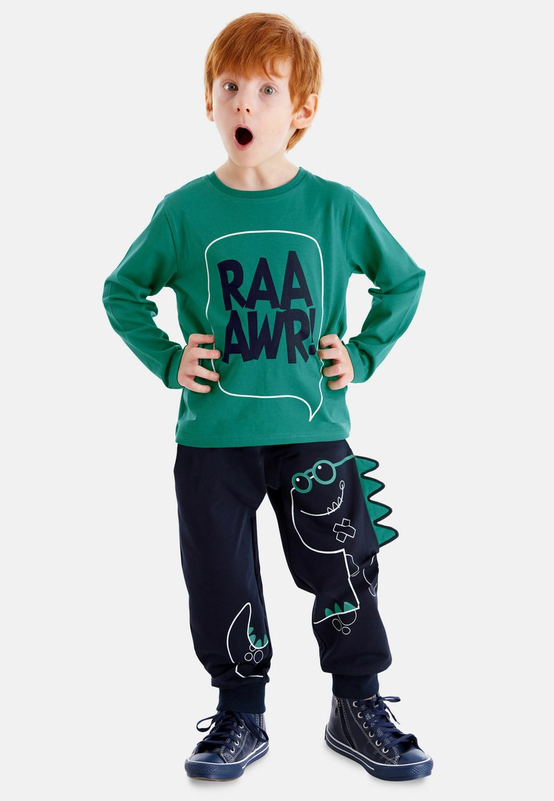 Camicia verde a maniche lunghe con stampa "RAAWR!", abbinata a pantaloni della tuta blu scuri con un design di dinosauro e spine verdi; sneakers blu.