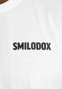 Valkoinen t-paita pyöreällä kauluksella, jossa on musta, kohotettu teksti "SMILODOX" rinnan kohdalla. Kangas näyttää pehmeältä ja sileältä.