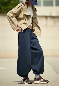 béžový skrátený trench coat, biela priliehajúca košeľa, tmavomodré voľné tepláky a hnedé bežecké topánky s bielymi akcentmi.