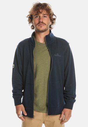 Navy zip-up jacket made of soft fabric with long sleeves, ribbed cuffs, and front pockets; layered over an olive green shirt.