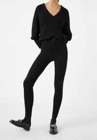 Jersey negro de punto con escote en V, combinado con unos jeans ajustados negros de cintura alta. El calzado presenta un material negro brillante con acentos de cadena dorada.